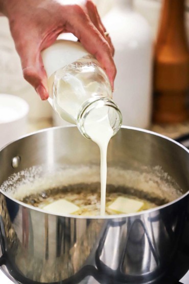 A person pouring heavy cream from a small milk jug into a silver saucepan filled with bubbling sugar water and melting butter.