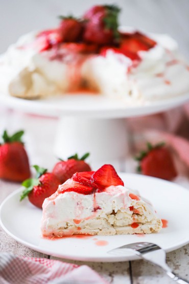 A slice of strawberry pavlova on a white dessert plate with the pavlova sitting on a cake stand in the background.