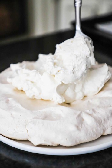 A person using a large spoon to add a layer of homemade whipped cream on top of a layer of baked meringue.