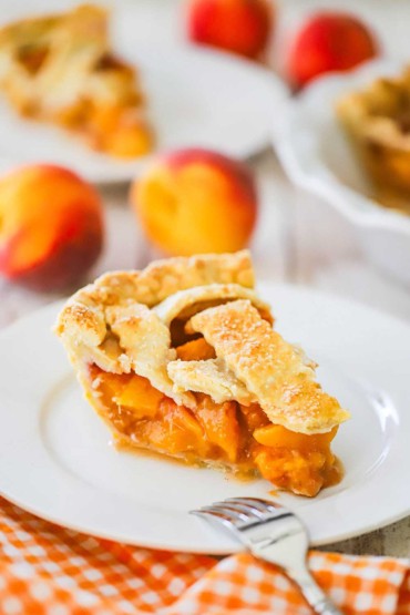 A slice of homemade peach pie sitting on a white dessert plate with whole fresh peaches sitting behind the plate.