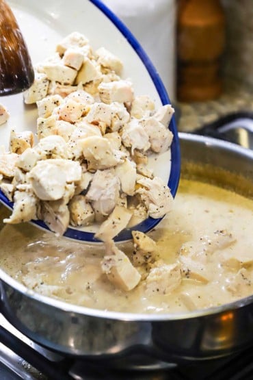 A person using a wooden spatula to transfer cooked strips of chicken into a skillet filled with a creamy sauce.