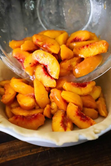 A person transferring slices of fresh peaches tossed in a sugar coating from a glass bowl into a pastry-lined pie dish.