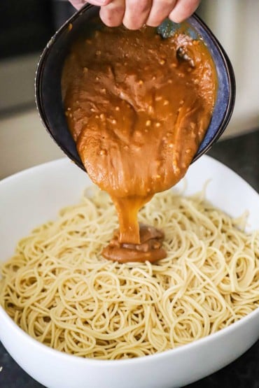A person pouring a peanut sauce from a blue bowl into a large white pasta bowl filled with cooked lo mein noodles.