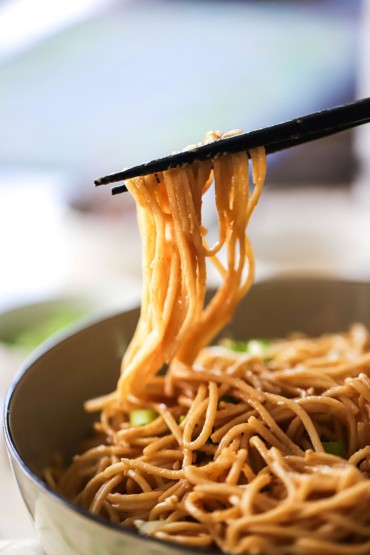 A person using a pair of chopsticks to lift up strands of cooked lo mein in a peanut sauce from a bowl.