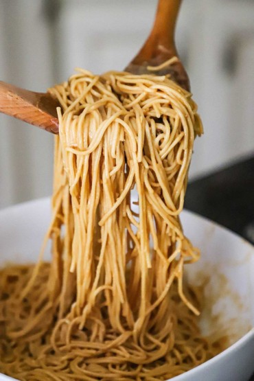 A person using two large wooden spoons to lift up cooked noodles that have coated with a peanut sauce.