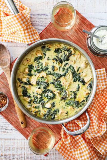 An overhead view of a large silver skillet filled with creamy chicken Florentine with a wooden spoon and small bowl of Parmesan cheese sitting nearby.
