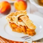 A slice of homemade peach pie sitting on a white dessert plate with whole fresh peaches sitting behind the plate.