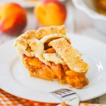 A slice of homemade peach pie sitting on a white dessert plate with whole fresh peaches sitting behind the plate.