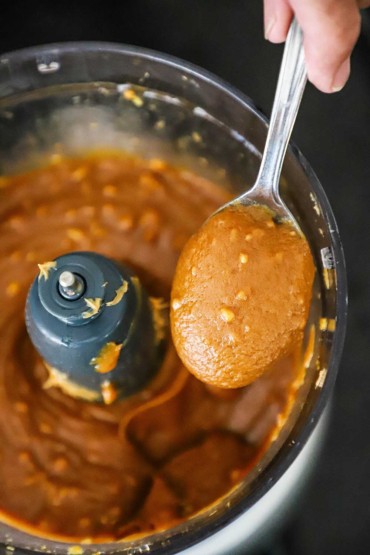 A person holding up a spoon filled with a peanut sauce from a food processor.