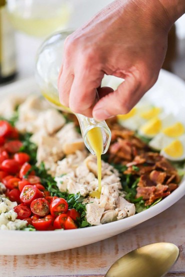 A person pouring a light-colored vinaigrette from a glass salad dressing holder over a platter of a prepared Cobb salad.