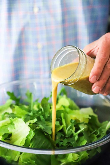 A person pouring a blue cheese vinaigrette from a small glass jar into a glass bowl filled with chopped Romaine lettuce and baby arugula.
