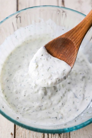 A person holding a wooden spoon filled with homemade tzatziki sauce over a glass bowl filled with the sauce.