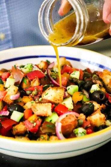 A person pouring a red wine vinaigrette over a bowl filled with croutons, chopped tomatoes, black olives, cubed cucumbers, and sliced red onions.