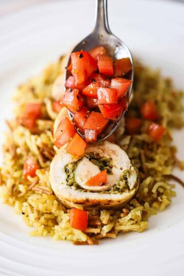 A person using a spoon to transfer a chopped tomato relish over slice pieces of cooked rolled up chicken that's been stuffed with pesto sauce.