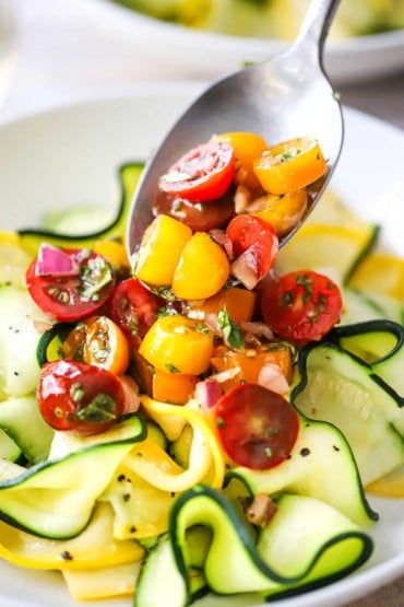 A person spooning a tri-color cherry tomato relish on top of thin slices of softened zucchini and yellow squash in a white dinner bowl.