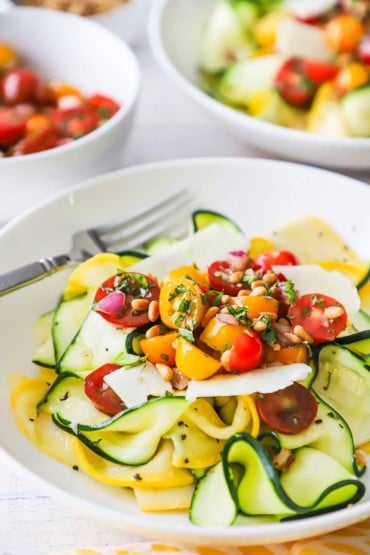A straight-on view of a bowl filled with a zucchini and yellow squash salad with tomato relish and fresh Parmesan shavings.