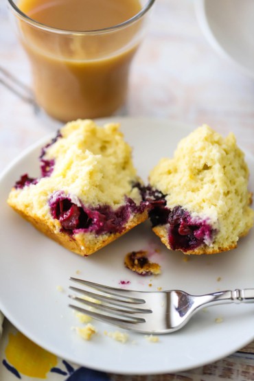 A straight-on view of a lemon blueberry muffin that has been split in half on a small white plate sitting next to a glass mug full of coffee.