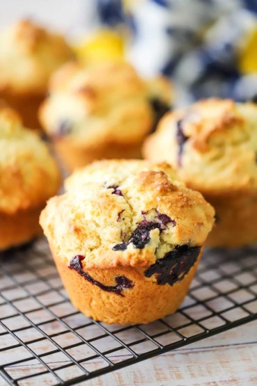 A straight-on view of a lemon blueberry jumbo muffin sitting on a baking rack with other muffins in the background.