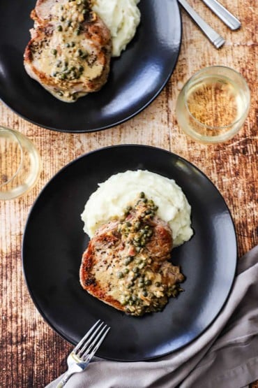 An overhead view of two black plates filled with a pile of mashed potatoes and a seared pork chop with caper sauce poured over the top.