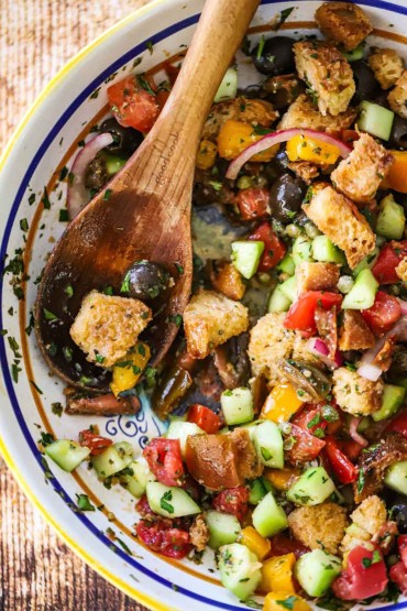 An overhead view a past bowl filled with a tomato and bread salad that has had some of it removed with a large wooden spoon that inserted into the side of the salad.