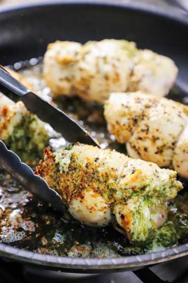 A person using a pair of metal tongs to turn a rolled up piece of chicken that is stuffed with pesto sauce and is being seared in a skillet.