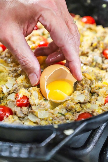 A person transferring a whole egg from a cracked shell into a skillet filled with a mixture of cooked sausage, tomatoes, and cheese.