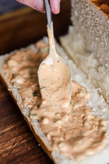 A person transferring Cajun remoulade from a large silver spoon onto the lower half of an open French roll.