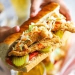 A person holding a large fried catfish po-boy that is stuffed with crispy fillet of catfish, coleslaw, hot sauce, pickles, and tomatoes.