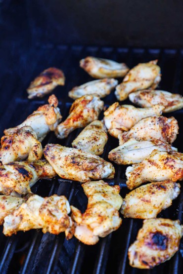 About twenty seasoned chicken wings cooking on the grates of a gas gril.