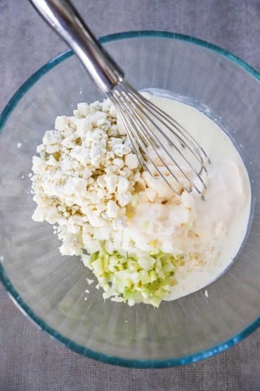 An overhead view of a medium-sized glass bowl holding crumbled gorgonzola cheese, diced celery and onion, mayonnaise, and cream.