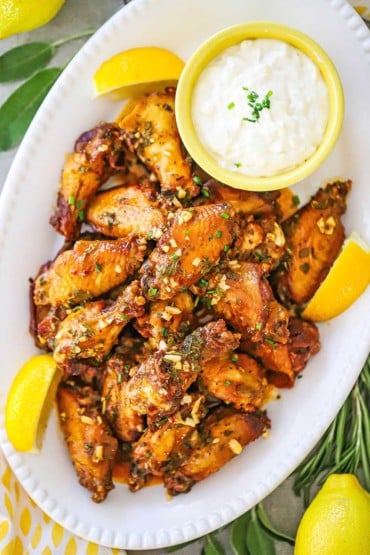 An overhead view of Italian-style wings on a platter along with a bowl of gorgonzola dipping sauce and lemon wedges.