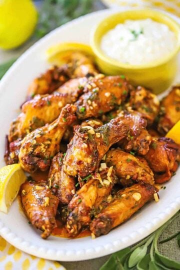 A straight-on view of Italian-style wings stacked onto a white oval platter with a yellow bowl of gorgonzola dipping sauce nearby.