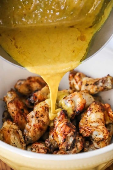A yellow curry coconut sauce being transferred from a silver saucepan into a bowl filled with grilled party wings.