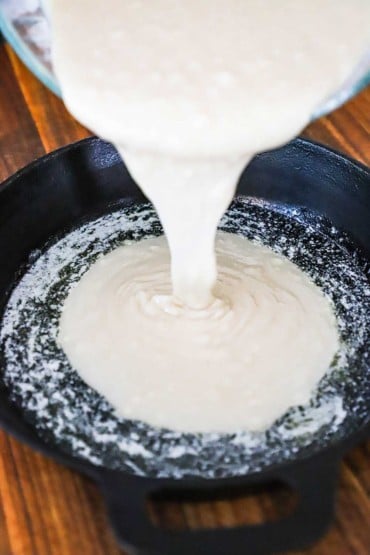 A thick creamy batter being poured into a cast-iron skillet that has a layer of melted butter in it.