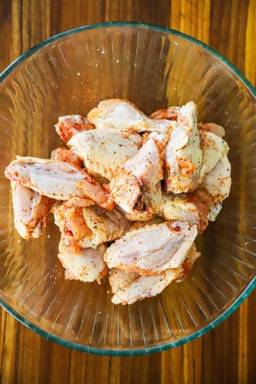 An overhead view of a large glass bowl filled with uncooked chicken wings that have been seasoned with salt, pepper, and paprika.