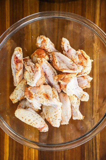 An overhead view of uncooked chicken wings in a glass bowl that have been seasoned with salt, pepper, and paprika.