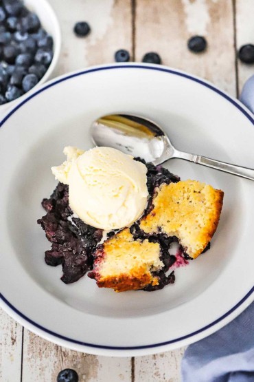 An overhead view of a white serving bowl filled with a helping of blueberry cobbler topped with a scoop of vanilla ice cream with a spoon off to the side.