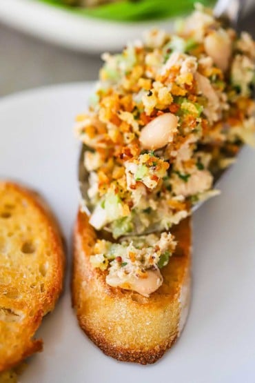 A person using a large metal spoon to transfer an albacore tuna and white bean salad on top of a toasted crostini slice of bread.