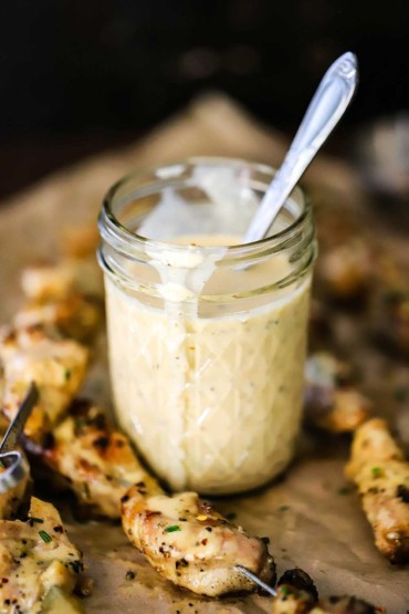A straight-on view of a small glass jar filled with Alabama white sauce next to grilled chicken on metal skewers.