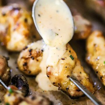 Alabama white sauce transferring onto a piece of skewered grilled chicken from a large silver spoon.