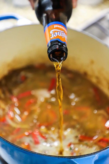 A person pouring beer from a brown bottle into a large Dutch oven filled with broth and sautéed onions and red bell peppers.
