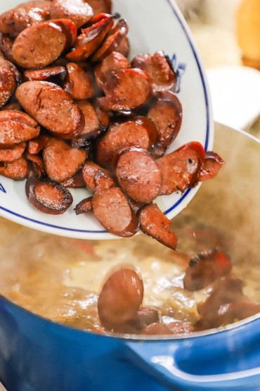 A person dumping sautéed medallions of kielbasa sausage into a Dutch oven from an oval platter.