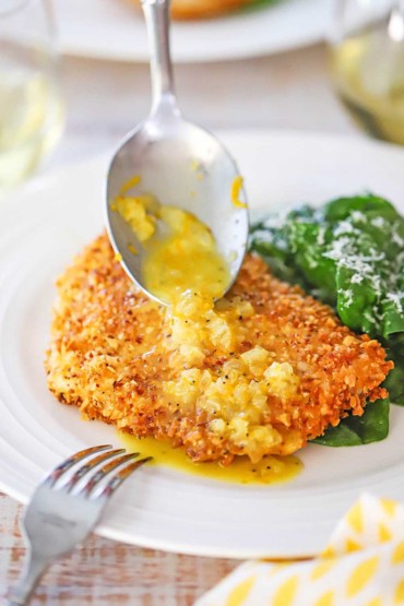 A dinner spoon being used to drizzle an orange shallot sauce over an almond crusted fillet of cod next to a spinach salad.