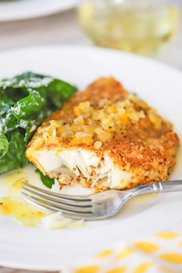 A close-up view of a fillet of almond crusted cod with orange sauce that has bite taken out of it and is on a white dinner plate.