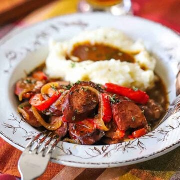 A close-up view of an antique bowl filled with a helping of beer-braised kielbasa and peppers next to a pile of mashed potatoes and gravy.