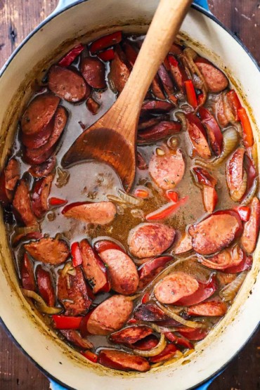 An overhead view of an oval Dutch oven filled with beer-braised kielbasa and onions with a large wooden spoon inserted into it.