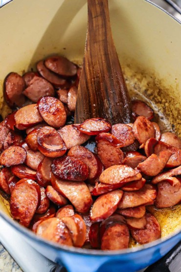 A close-up view of medallions of kielbasa sausage that are being sautéed in an oval Dutch oven with a wooden spatula.