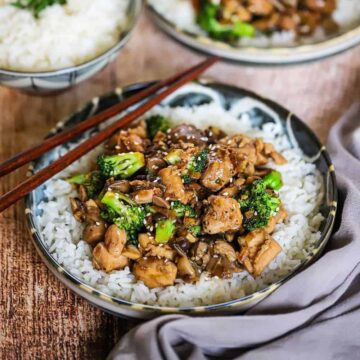 A close-up view of a shallow bowl filled with a layer of white rice topped with chicken and broccoli stir-fry with mushrooms.
