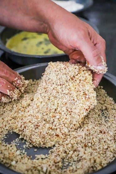 A person holding up an uncooked fillet of cod that is in a baking pan filled with a breading of pulverized almonds, Parmesan cheese, and breadcrumbes.