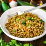 A straight-on view of a white serving bowl filled with Cajun dirty rice topped with chopped scallions and several dashes of cayenne hot sauce.